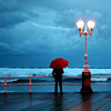revolutions: A person with a red umbrella standing under a streetlamp by the seafront (beside the seaside)