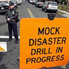john: Orange warning sign on street. Sign reads "MOCK DISASTER DRILL IN PROGRESS" (mock: disaster drill in progress)