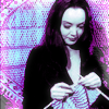 auroracloud: a dark-haired woman (Morticia Addams) knitting and smiling, in purple colour scheme (crafty)