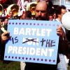 writer_at_the_table: a screenshot from The West Wing of Toby Ziegler holding a Bartlet for President campaign sign with "for" crossed out and "is the" written in marker (Bartlet is the President)