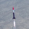 emceeaich: A purple and pink model rocket under thrust accellerates upwards, with a distant mountain in the background (high power rocketry)