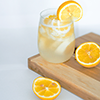 enemytosleep: a clear glass is full of a summer-time beverage with several slices of orange fruit within and around the glass. It all sits on a wooden table or board with a light-colored background (Sunshine)