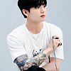 celli: Jungkook from BTS, in a white shirt, with the tattoos on his arms showing as he clicks the button to take a selfie (JK selfie)