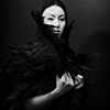 ivoryandhorn: A black and white photo of a woman against a black background, wearing a black feathery cape. Her pale face and hands stand out starkly against the black. (Default)