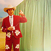 militarypenguin: Jupe, clad in a red cowboy suit and white hat, standing in front of a faint green sheet concealing something large and rectangular, from Nope. (Nope - you'll leave here different)