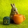 coinin: Photo of a small brown bunny figure drinking tea, against a bright green background. (Default)