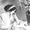 inchoatewords: A woman sitting at a table writing. She is leaning her head on her other hand and looking down at the paper. She is dressed in a light colored dress and has dark hair swept back with a light colored hairband. There is a large vase of flowers in the right of the picture. (writing)