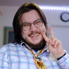 brennanbrown: A person with long dark hair, glasses, and a beard grins broadly while holding up a peace sign. They wear a light blue plaid button-up shirt and a beaded medallion necklace featuring a thunderbird design with feather and claw pendants. Colourful Buddhist prayer flags hang in the background. (Default)