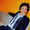 crossedwires: rumpled john cho sitting down and leaning back against a bright orange wall, smiling at something out of frame (john cho rumpled)