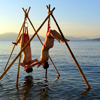thingswithwings: Two people in upside-down rope suspension hanging from wooden tripods above the ocean; they're reaching out their arms t (queer - suspended over the ocean)