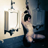thingswithwings: A naked person squatting, chained up, between two urinals, as if they are a urinal themselves. (queer - chained up as a urinal)