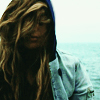 binz: a woman with long blond hair has her sweatshirt hood up and her hair over her eyes and stands in front of the ocean. ([ stock ] wavewashed)