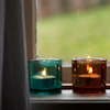 jules_robin: Two votive candles sitting on a windowsill, one in a teal glass the other in a brown glass. (Candles in the window)