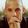 thirteen_pillars: An olive-skinned bald man (Vin Diesel) looks at the camera. His hands are linked and propped in front of his mouth. (Default)