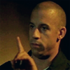 thirteen_pillars: An olive skinned bald man (Vin Diesel) holds up a finger, telling someone off camera to pause or be quiet. (Hush)