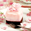 milkymoon: A pink petit-four sitting on a plate with flowers on it, with a fork next to it. It's decorated with small pink flowers. (Flower cake.)